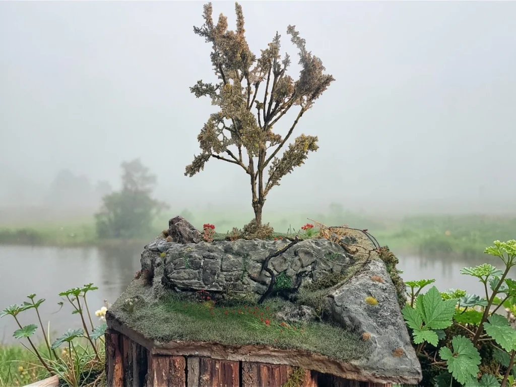 Diorama paesaggistico con muro in pietra a secco, albero alto, vegetazione e prato fiorito