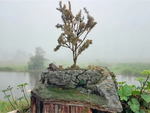 Diorama paesaggistico con muro in pietra a secco, albero alto, vegetazione e prato fiorito