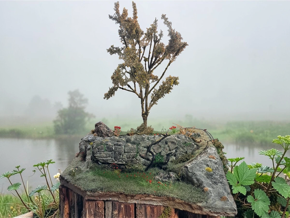 Diorama paesaggistico con muro in pietra a secco, albero alto, vegetazione e prato fiorito