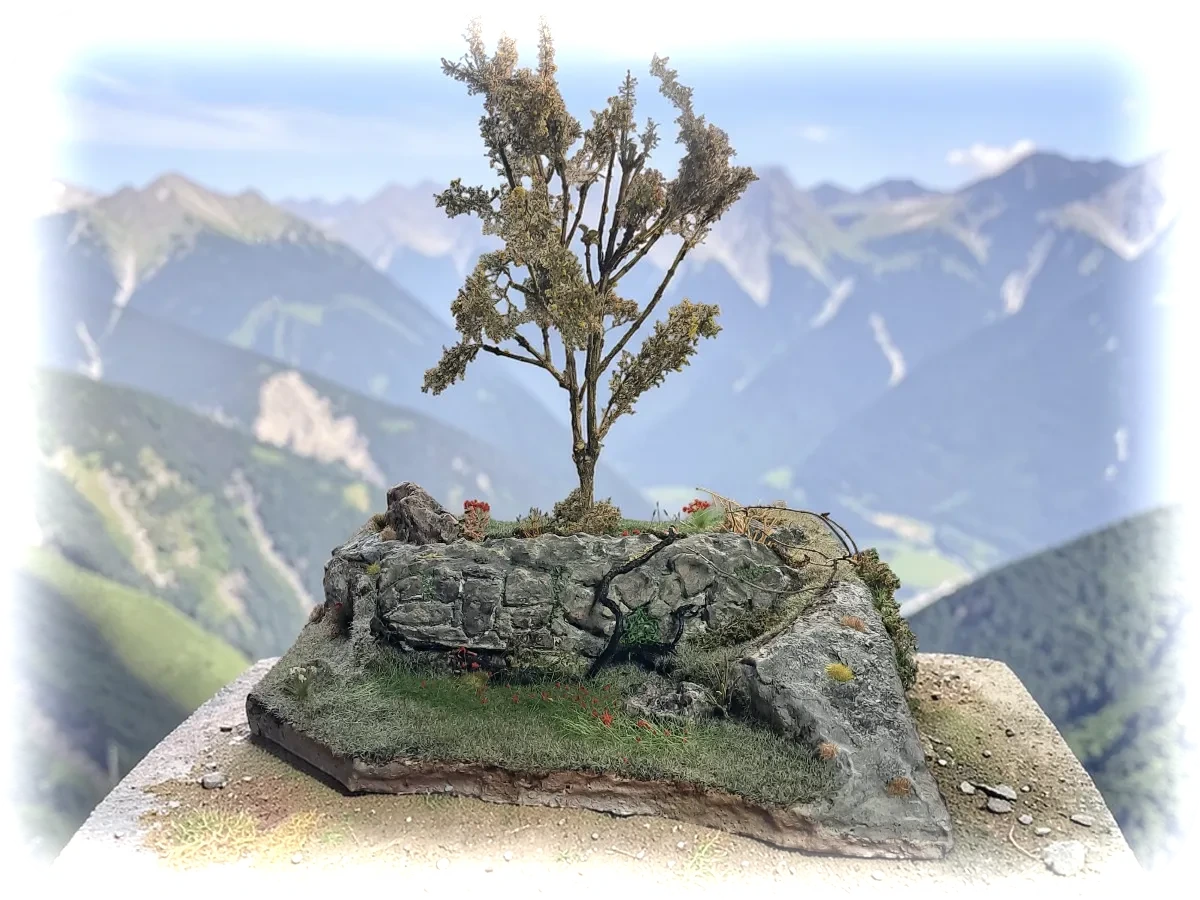 Diorama paesaggistico con muro in pietra a secco, albero alto, vegetazione e prato fiorito